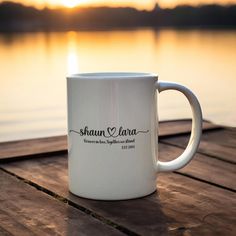 a white coffee mug sitting on top of a wooden table next to a lake at sunset