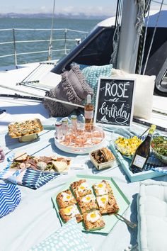 a table with food and drinks on it next to the ocean in front of a boat