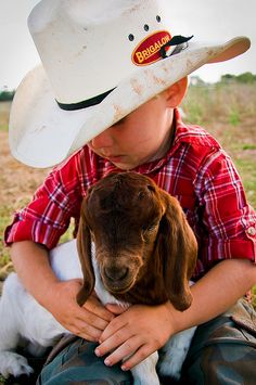 a young boy wearing a cowboy hat holding a baby goat in his lap while sitting on the ground