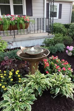 a bird bath in the middle of a flower bed with flowers around it and a house in the background