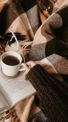 a person is reading a book and holding a cup of coffee while laying on a plaid blanket