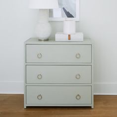 a white lamp sitting on top of a dresser next to a painting and a vase