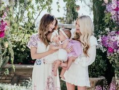 three women are holding two baby girls in front of a flower covered arbor with pink and purple flowers