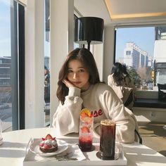 a woman sitting at a table in front of a window with drinks and food on it