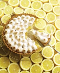 a lemon tart with a slice missing from it on a glass plate surrounded by sliced lemons