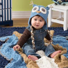 a baby sitting on a blue rug wearing a knitted owl hat and overalls