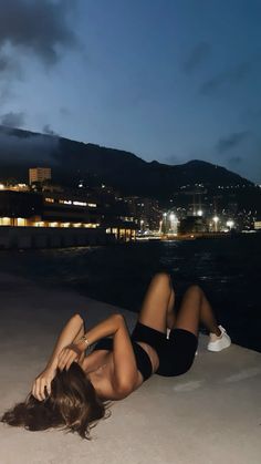 a woman laying on the ground in front of water at night with buildings and lights behind her