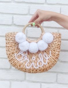 a hand holding a straw bag with pom - poms on the handle and white lettering