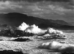 black and white photograph of waves crashing on the rocks in front of an island with mountains