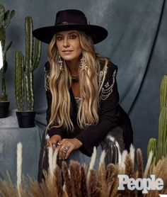 a woman with long blonde hair wearing a black hat sitting in front of cactuses