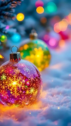 two christmas ornaments sitting in the snow next to a tree with colorful lights on it