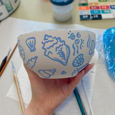 a hand holding a bowl with blue designs on it next to paintbrushes and water bottles