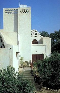 a white building with an arched doorway and stairs leading up to the front door is surrounded by greenery