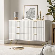 a white dresser with gold handles in a room next to a potted plant and a book