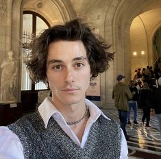 a young man with curly hair wearing a white shirt and black vest standing in an ornate building