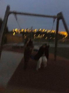 two people are playing on a swing set at night with the lights in the background