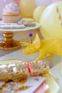 a yellow dinosaur figurine sitting on top of a table next to cupcakes