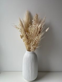 a white vase filled with dried plants on top of a table next to a wall