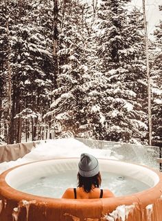 a woman sitting in an outdoor hot tub surrounded by snow covered trees and evergreens