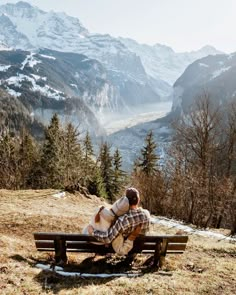 a man sitting on top of a wooden bench next to a forest filled with snow covered mountains