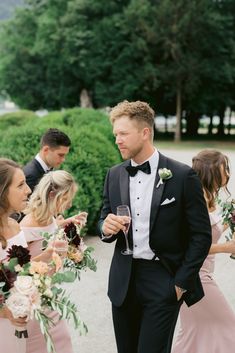 a man in a tuxedo standing next to other people holding wine glasses and flowers