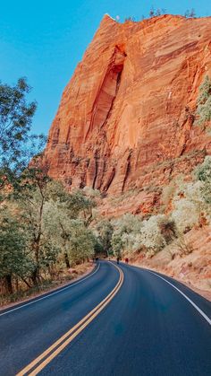 an empty road in front of a large rock formation with trees and bushes on both sides