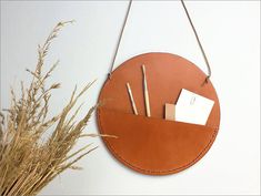 a round leather wall hanging with two pens and pencils in the pocket, next to some dried grass