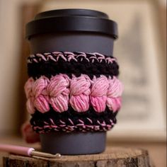 a coffee cup holder made out of crocheted yarn and a pink pen sitting on top of a wooden table