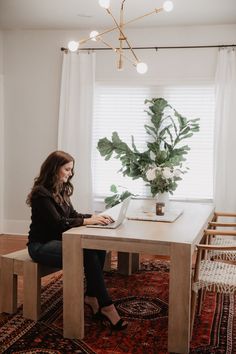 a woman sitting at a table with a laptop on her lap and potted plant in the window