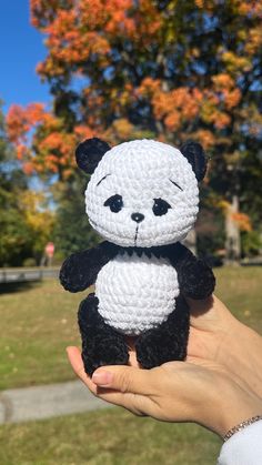 a hand holding a small crocheted panda bear in front of a tree with orange leaves