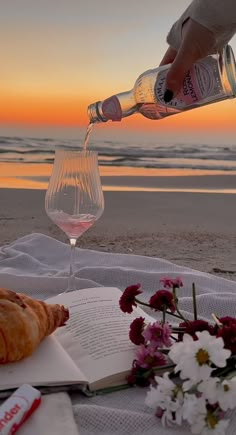 a person is pouring wine into a glass on the beach with flowers and croissants