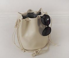 This unique bucket bag features soft, off-white faux (fake) leather, cotton rope shoulder strap, and a drawstring closure. You can tie the cotton rope to wear this bucket bag long or short. Details:-Dimensions when laid flat: 9" length (top to bottom), 7.5" diameter round bottom-Shoulder strap measures approx. 45-50" total-Soft, off-white faux (fake) leather-Solid light color interior lining-No internal pocketBecause each bag is made by hand, the measurement is approximate and may vary slightly Spring Travel Cream Bucket Bag, Cream Bucket Bag With Leather Handles For Spring, Spring Cream Bucket Bag With Leather Handles, Casual Cream Bucket Bag For Daily Use, Cream Bucket Bag With Dust Bag For Daily Use, Casual Cream Bucket Bag Tote, Casual Everyday Bucket Drawstring Bag, Summer Cream Bucket Bag, Spring Cream Bucket Bag With Braided Handles