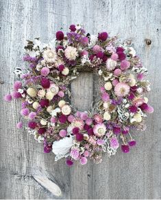 a wreath with purple and white flowers hanging on a wooden door frame, in front of a weathered wall