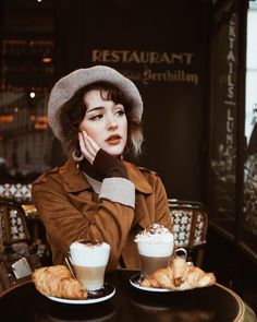 a woman sitting at a table with two cups of coffee and croissants