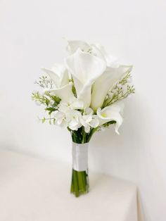 white flowers are arranged in a vase on a table with a white wall behind it