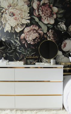 a white dresser sitting on top of a bed next to a wall covered in flowers