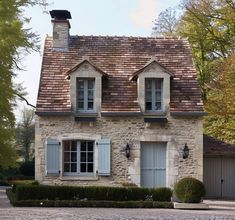 an old stone house with two windows and shutters on the front, surrounded by greenery