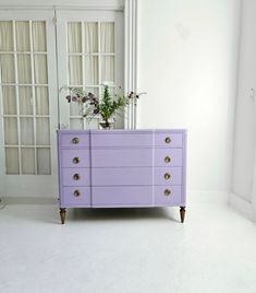 a purple dresser with flowers on top in front of a white wall and glass doors