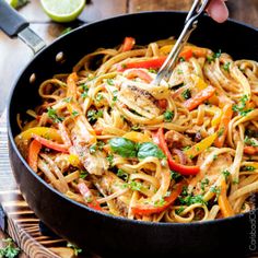 a pan filled with pasta and vegetables on top of a wooden table next to limes