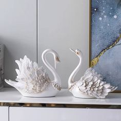 two white swans sitting on top of a counter next to a painting and a vase