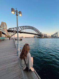 a woman sitting on a pier looking out at the water and a bridge in the background