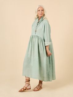 an older woman in a green dress standing on a beige background with her hands behind her back