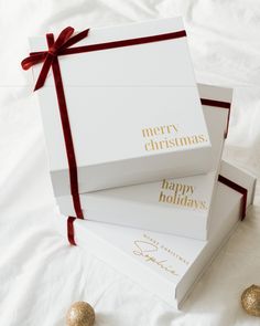 two white boxes with red ribbon and gold foil lettering on them sitting on a bed