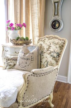 a chair with pillows and flowers on it in front of a window, near a clock