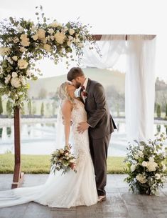 a bride and groom kissing under an arch decorated with white flowers at their wedding ceremony