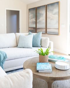 a living room with white couches and blue pillows on the coffee table in front of two paintings