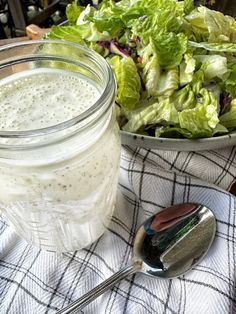 a salad in a jar next to a bowl of salad dressing and a spoon on a table