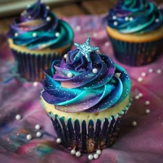 three cupcakes with blue, purple and green frosting on a pink table cloth