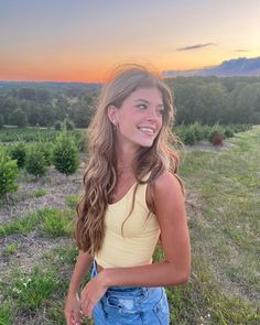 a beautiful young woman standing on top of a grass covered field next to a forest