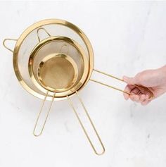 a person holding a metal strainer over a white counter top with gold colored handles